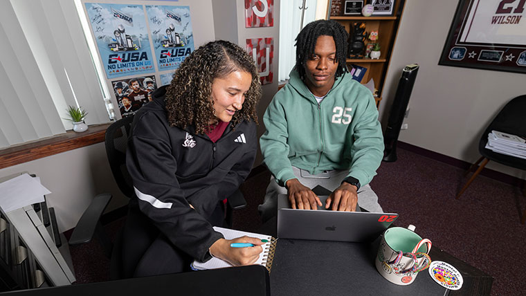 An advisor writes in a notebook while her student-athlete advisee types on a laptop, as they review his schedule.