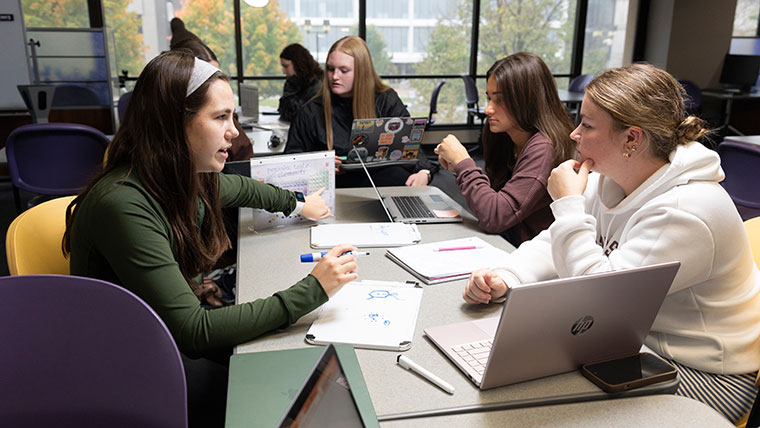 A Bear CLAW tutor points to a chemistry chart on a table. Three other students at the table are listening and studying.