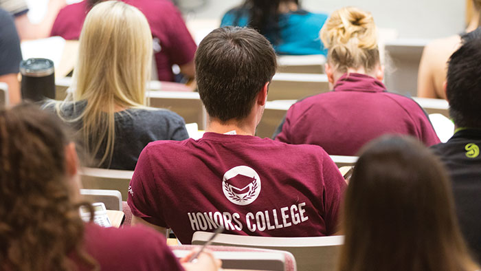 The back of students sitting in a lecture hall; the back of one student's shirt says "Honors College"