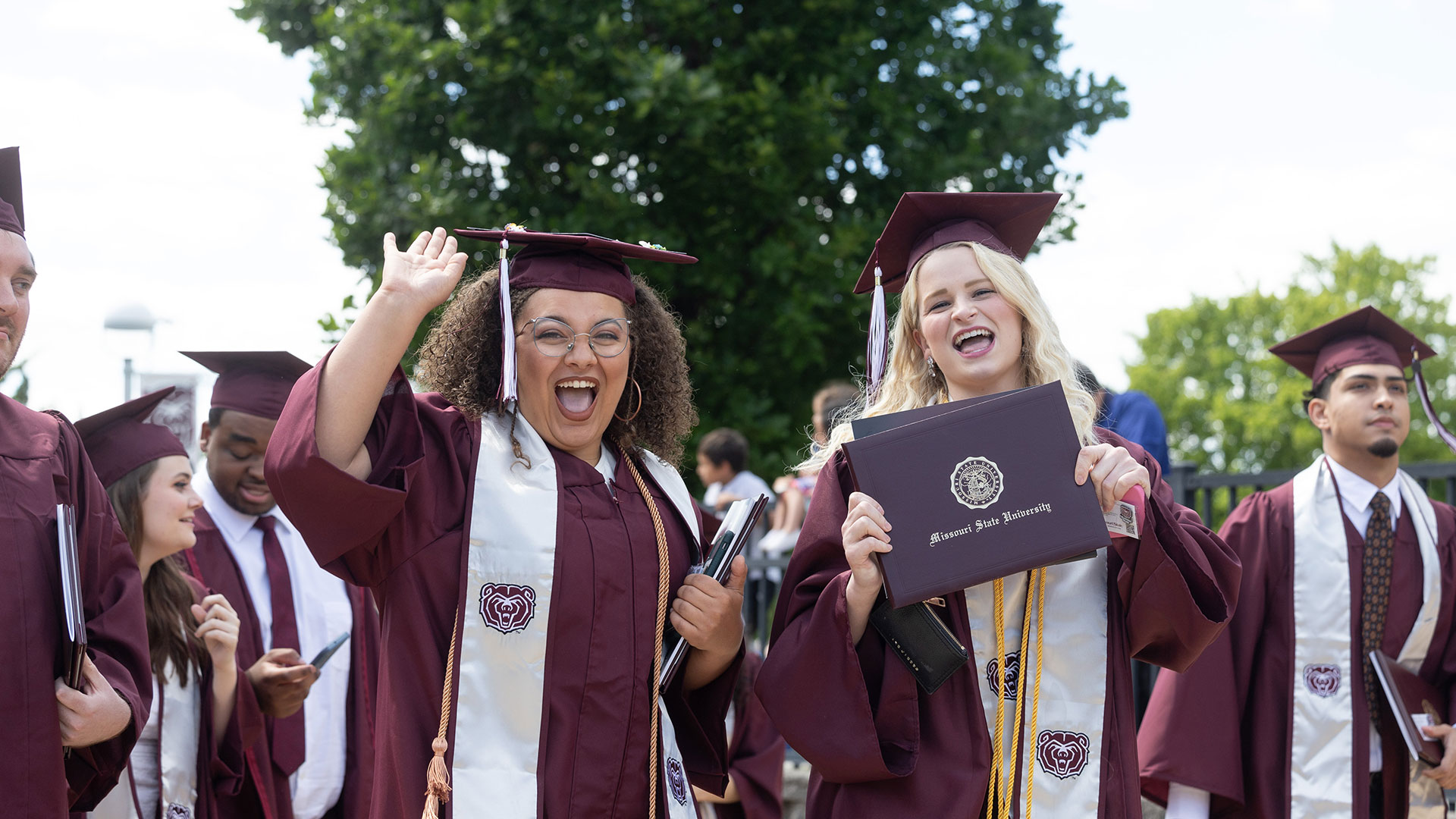 Two graduates smile and cheer, as one proudly displays her diploma.