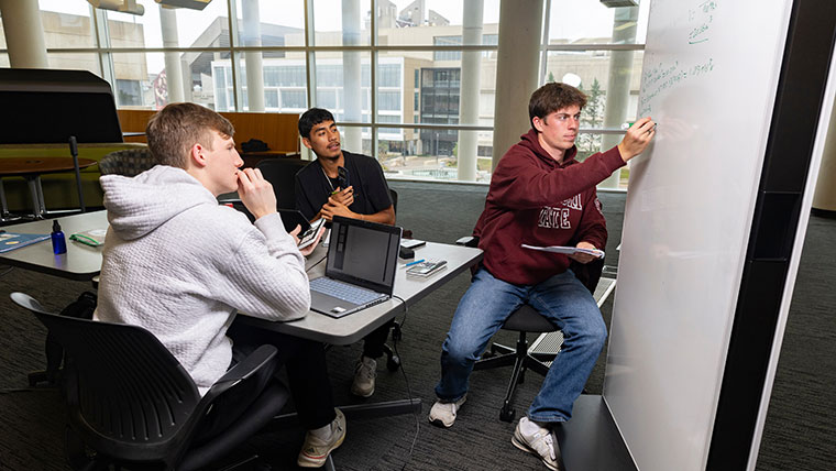 Three students study together in the Meyer Library. Two of the students watch as a third student writes on a white board.