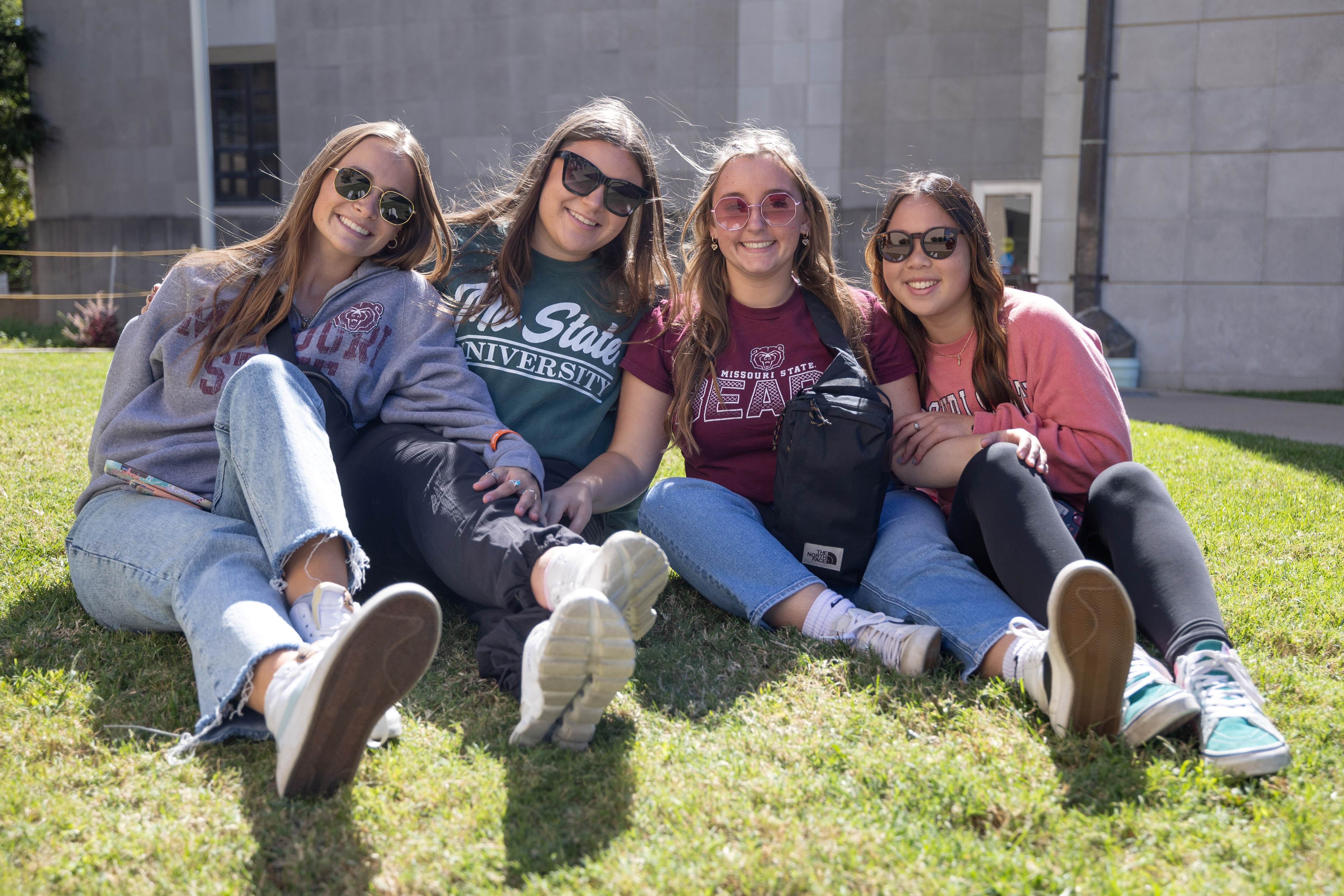 Four smiling female students in sunglasses and casual college apparel (gray, dark green, maroon, pink) sit on green grass on a sunny day. A large gray building is behind them.