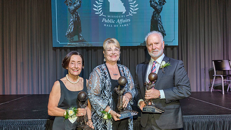Three smiling recipients holding their award in front of a stage with a Public Affairs Hall of Fame banner behind them.