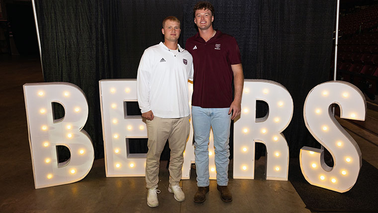 Two Missouri State athletes standing in front of a large lighted sign that says BEARS.