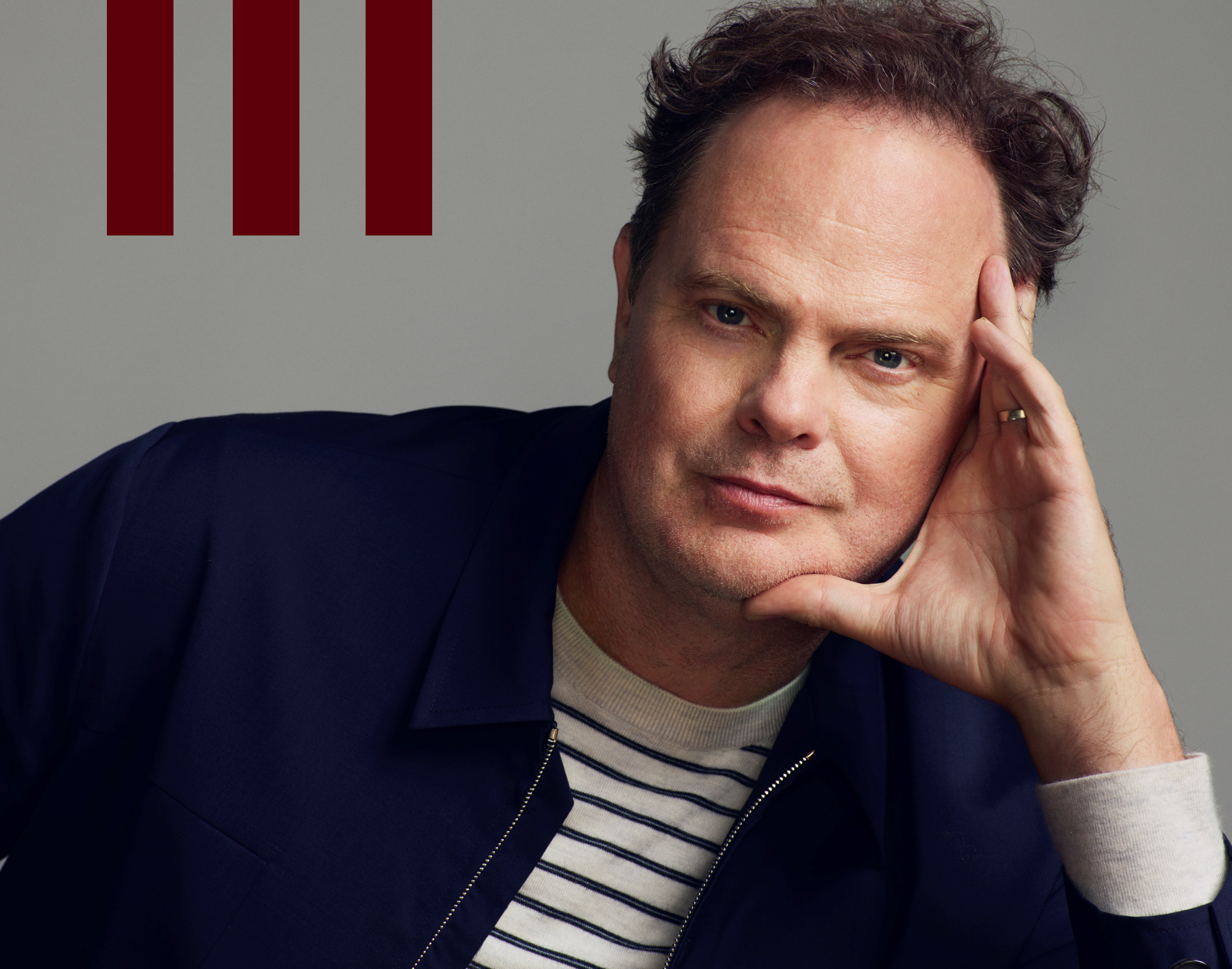 Man with blue eyes, dark wavy hair, & light stubble in a navy jacket & striped shirt, chin rested on hand, looking at camera. Grey background with three red bars top left.