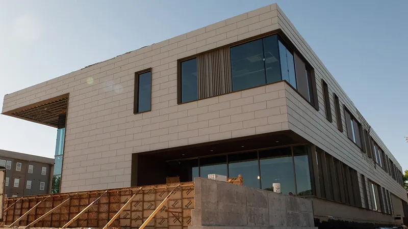 Modern building under construction: light beige block facade, dark-framed windows (one with vertical louvers), and an overhanging section on the left. Concrete forms in foreground, clear sky overhead.