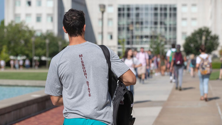 A student wearing a Missouri State T-shirt walks to Strong Hall on a sunny day.