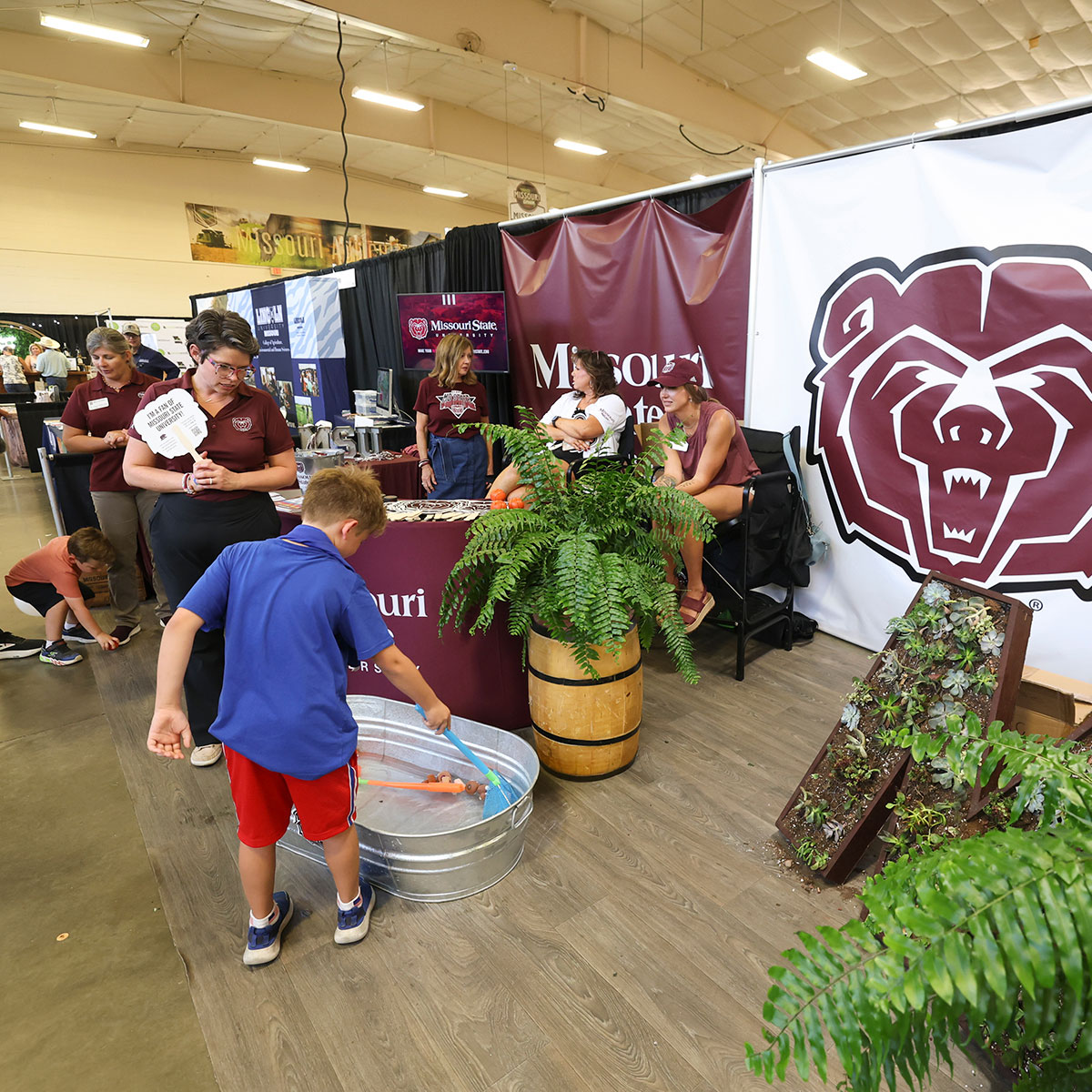 Mo States employees supervise the booth at the fair, as a child plays a game at the booth by attempting to scoop up a plastic duck from a tub of water.