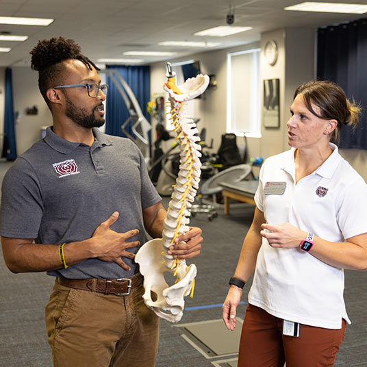 A physical therapy holds a model of a human spine while talking about techniques with a physical therapy professor.
