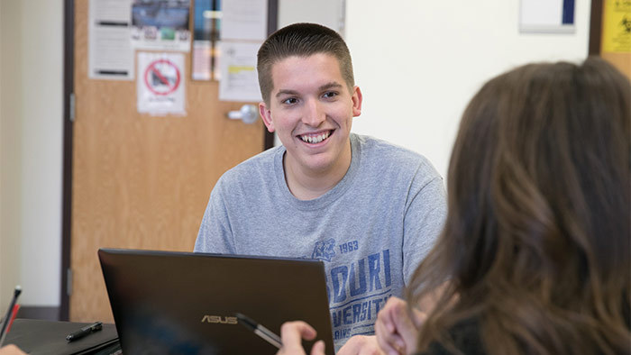 Student at a computer smiles across a desk at an advisor