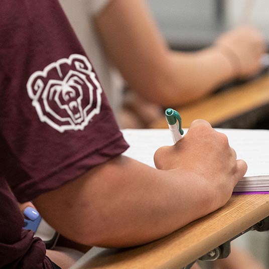 Student wearing a Bearwear shirt takes notes in class.