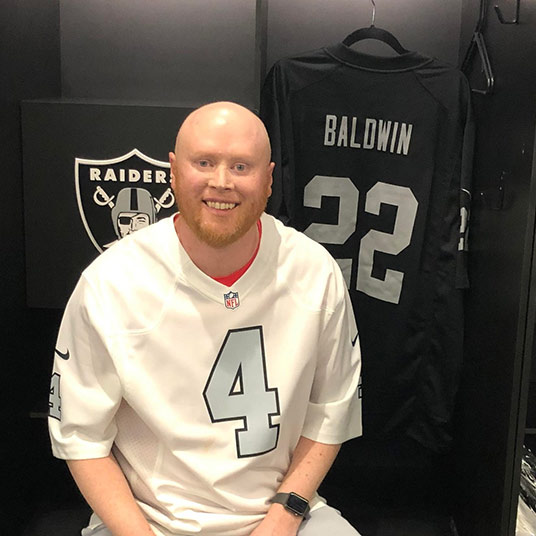 Missouri State alum Tyler Baldwin wearing a white Las Vegas Raiders jersey. A black Raiders jersey with the name Baldwin on it is hung up in the background.