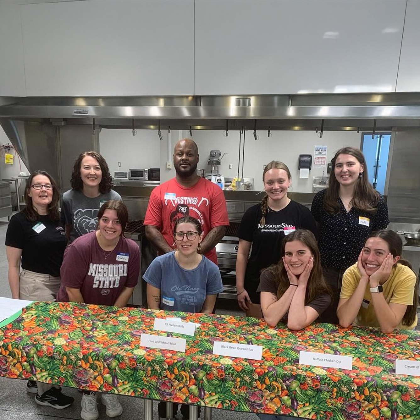 Dietetics students (the front row seated at a table and the back row standing behind them) have their class in a kitchen.