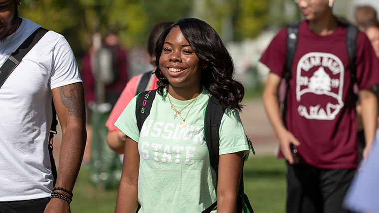 A student smiles at the camera while walking to class on a nice sunny day