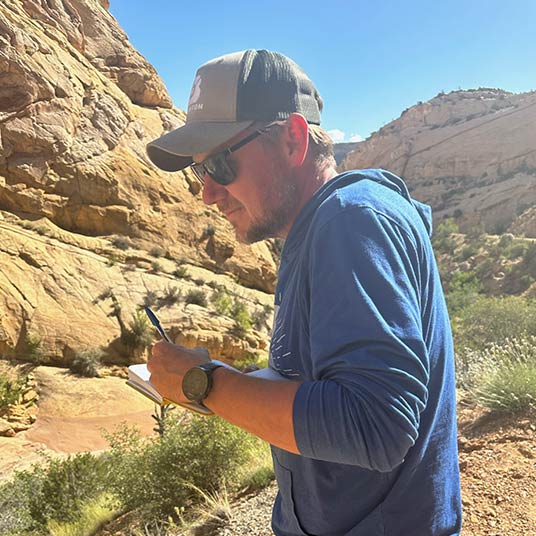 Dr. Matthew McKay, associate professor of geology, takes note of his observations in a notebook as he explores some rocky terrain.