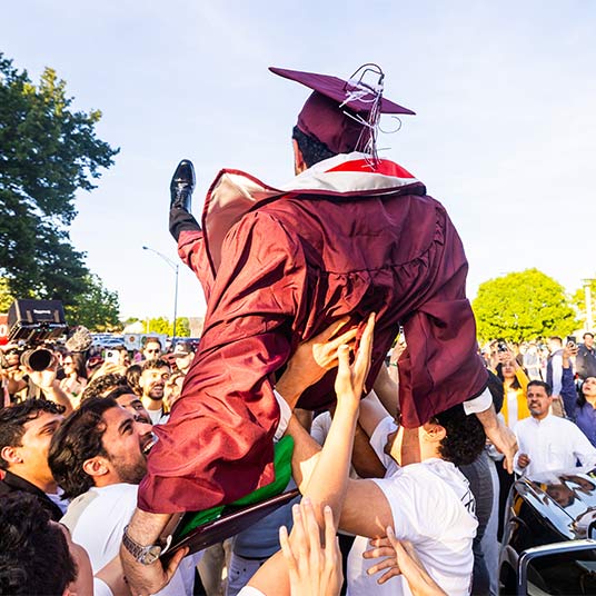 A student in commencement regalia is lifted in the air by his friends and family.
