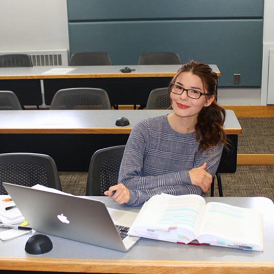 Sarah Lynne Skrzypczak, a Missouri State alumna with a degree in sociology, is seated in a classroom with her laptop and textbook open in front of her.