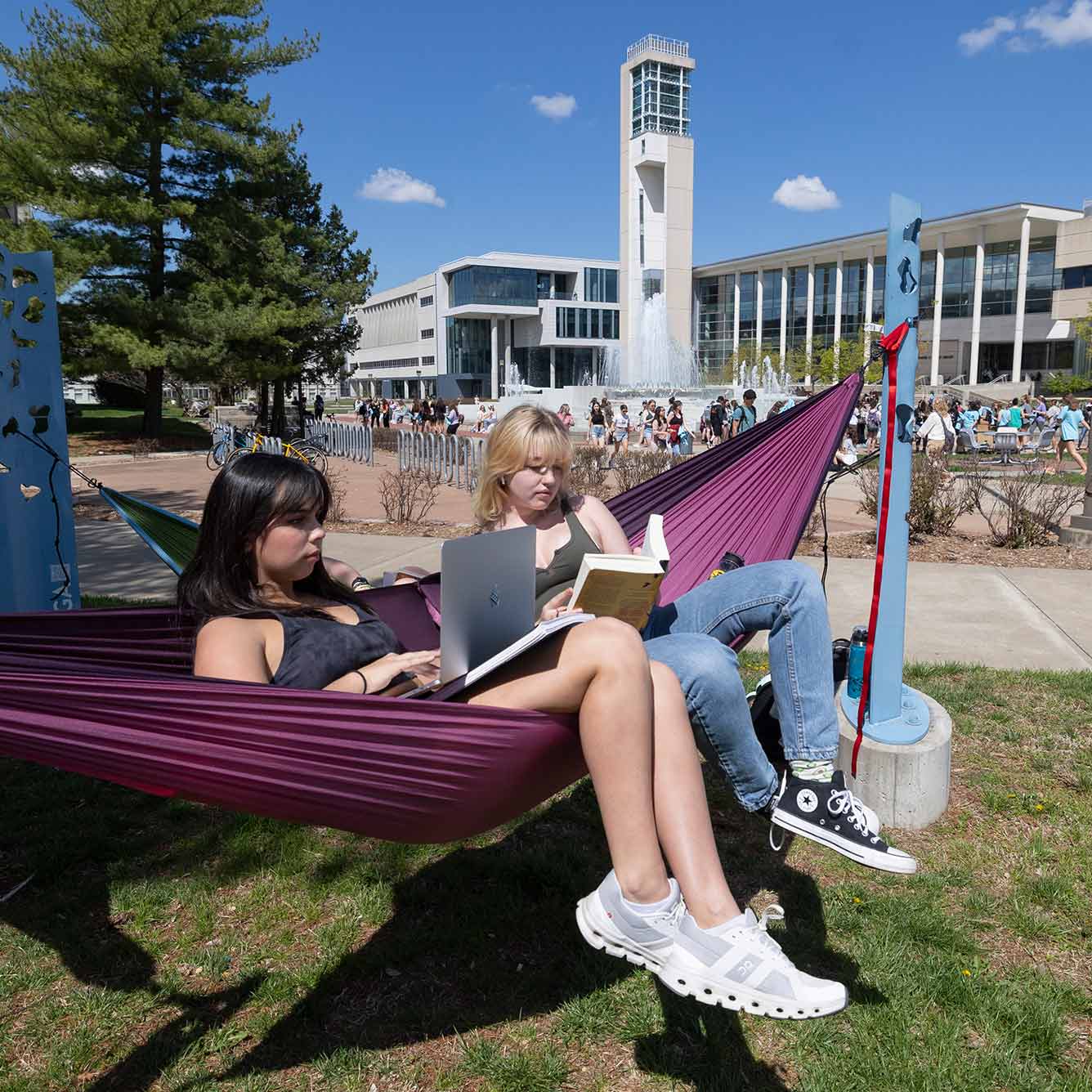 Two students sitting in a hammock on a sunny day while using their laptops. A group of students walk past the fountain in the background.