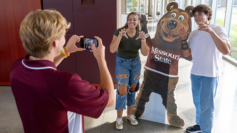 Students posing next to a cutout of Boomer at the Welcome Center as another student takes their photo.
