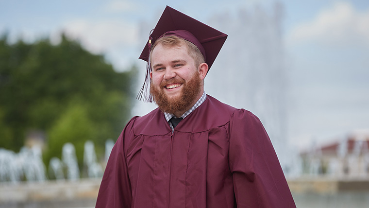 Smiling Missouri State graduate, in cap and gown, standing in front of John Q. Hammons Fountains. 