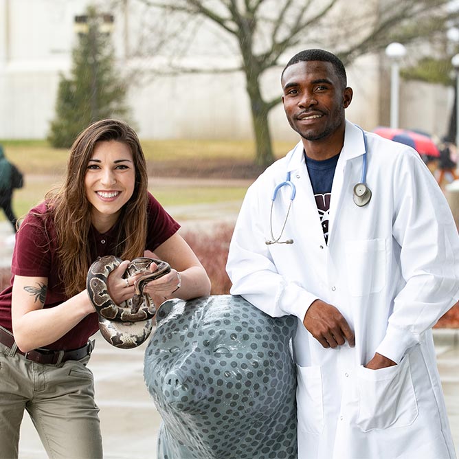 A student holding a snake next to a student in a white lab coat with stethoscope. 