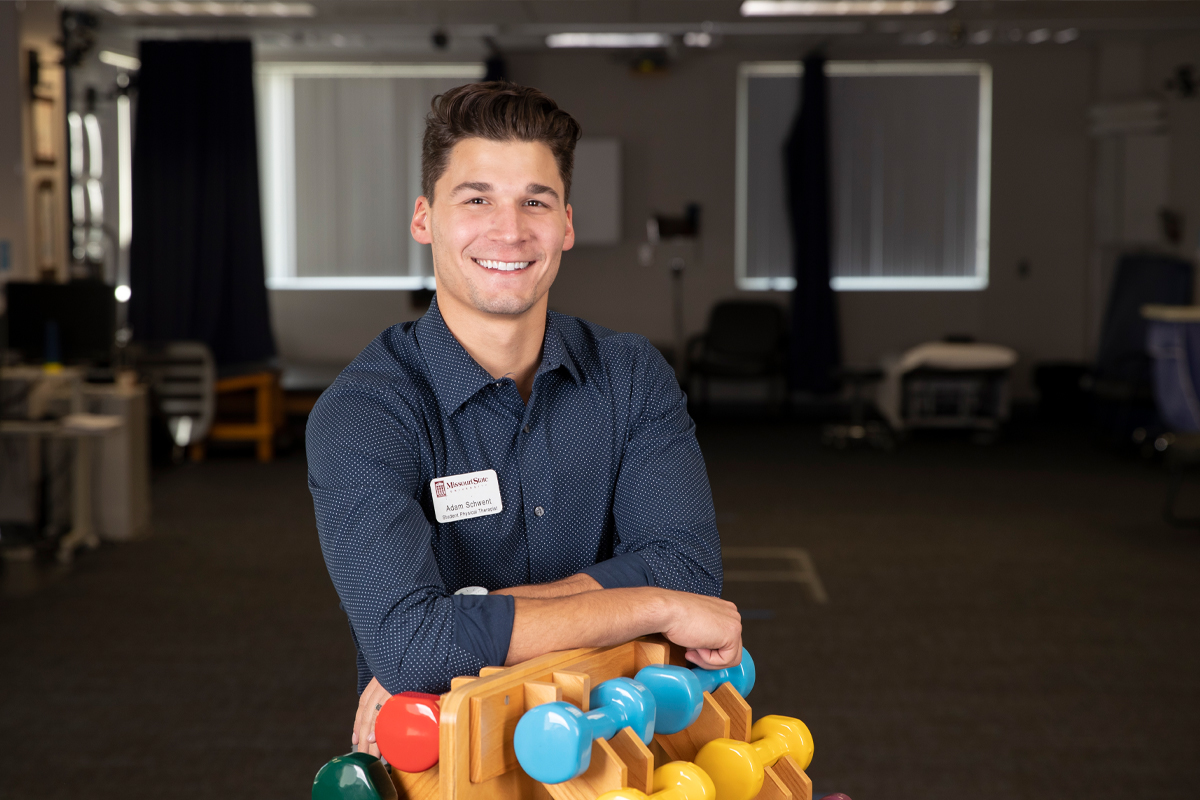 An occupational therapy student smiles while leaning on a dumb bell tower